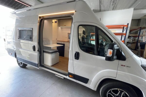 Perspectiva exterior de una Fiat Ducato L4H3 2+2 Garaje, con la puerta corredera abierta mostrando el interior equipado y el área de estar. La furgoneta está estacionada en un garaje con suficiente espacio alrededor, lo que indica su tamaño considerable y capacidad funcional.