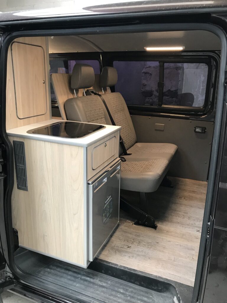 Detalles laterales (vista desde la puerta lateral) de los asientos traseros con cocina y nevera en la Camper Volkswagen Transporter T5 convertida por Baovan.