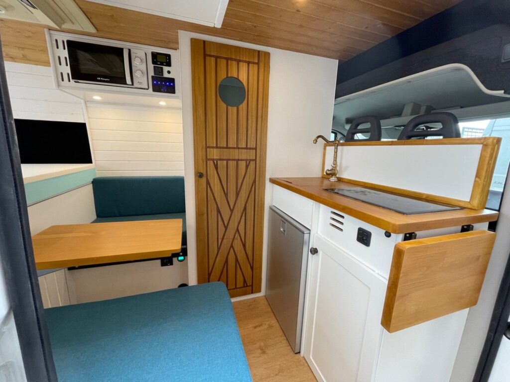 Detalhes da cozinha e área de descanso com pia, fogão, microondas e porta do banheiro, em acabamento branco e madeira, muito espaçoso na furgoneta Boxer L2H2 3+3