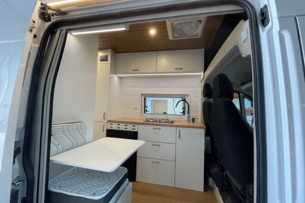 Interior de una Fiat Ducato L4H3 2+2 Garaje visto desde la puerta lateral abierta, mostrando un área de comedor con una mesa blanca desplegable y asiento con cojín de patrón geométrico. A la derecha, la cocina equipada con encimera de madera clara, fregadero con grifo negro, y armarios en tonos blancos y negros. Encima, armarios superiores blancos ofrecen almacenamiento adicional. La cabina del conductor se integra armoniosamente con el espacio habitable, y la iluminación es suave y acogedora, con una tira de luz LED en el techo.