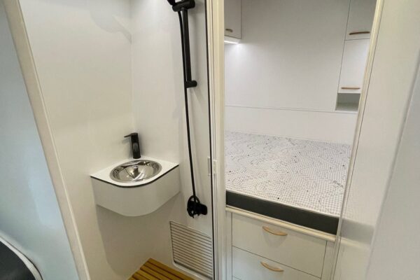 Vista interior del baño en una Fiat Ducato L4H3 2+2 Garaje, con una ducha con suelo de madera, lavabo moderno y ducha de mano negra. Se observa una claraboya que proporciona luz natural y un ambiente espacioso y limpio.