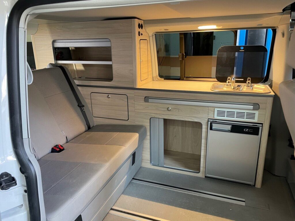 Cocina compacta en la Camper California T6 con mueble lateral, equipada con fregadero, grifo y refrigerador bajo la encimera.