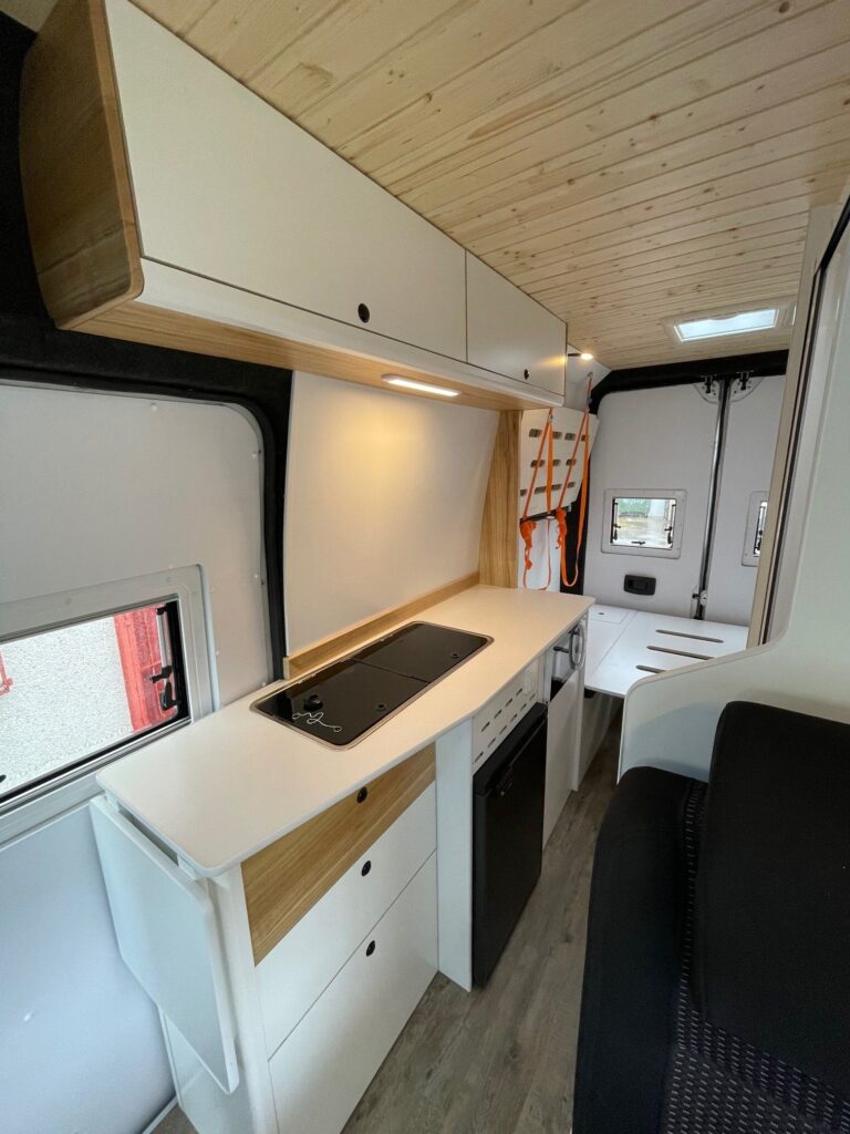 Detalle de la cocina, con fregadero, estufa, armarios, nevera y la cama al fondo en la Citroën Jumper L4H3 4+4, convertida por Baovan Camper.
