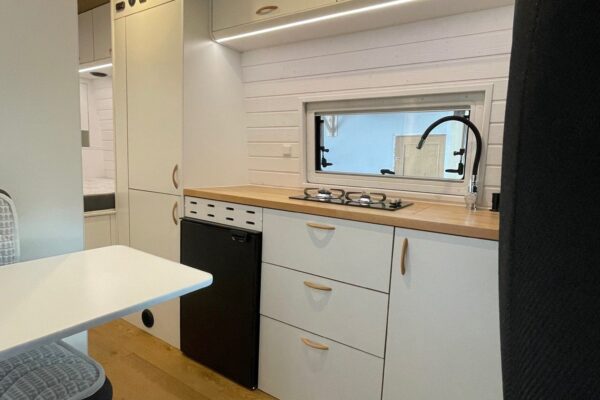 Vista de la cocina dentro de la Fiat Ducato L4H3 2+2, con una encimera de madera, fregadero y grifo negro, cajones blancos y una placa de cocina, junto a una mesa plegable y un asiento, todo bajo una iluminación suave.