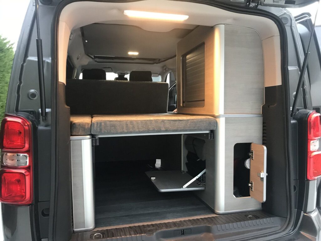 Vista externa de la Peugeot Traveller: puerta trasera abierta, armarios, cama montada, acabados tapizados