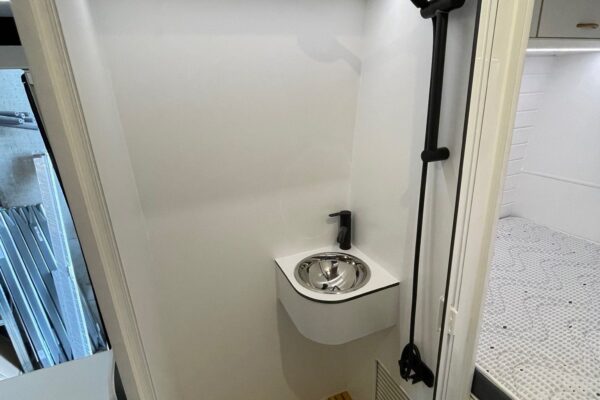Imagen del baño compacto en la Fiat Ducato L4H3 2+2, con una ducha con cabezal y grifería en negro, un pequeño lavamanos y suelo de madera, en un espacio optimizado con paredes blancas.