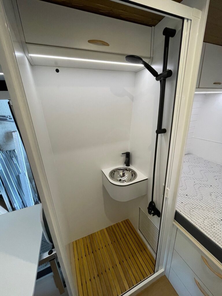 Imagen del baño compacto en la Fiat Ducato L4H3 2+2, con una ducha con cabezal y grifería en negro, un pequeño lavamanos y suelo de madera, en un espacio optimizado con paredes blancas.