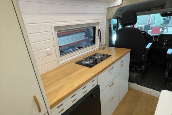 La cocina en la Fiat Ducato L4H3 2+2, mostrando la encimera y la placa de cocina desde una perspectiva que incluye el asiento del conductor, destacando la integración del espacio de vida con la cabina del conductor.
