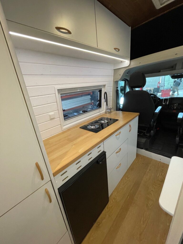 La cocina en la Fiat Ducato L4H3 2+2, mostrando la encimera y la placa de cocina desde una perspectiva que incluye el asiento del conductor, destacando la integración del espacio de vida con la cabina del conductor.