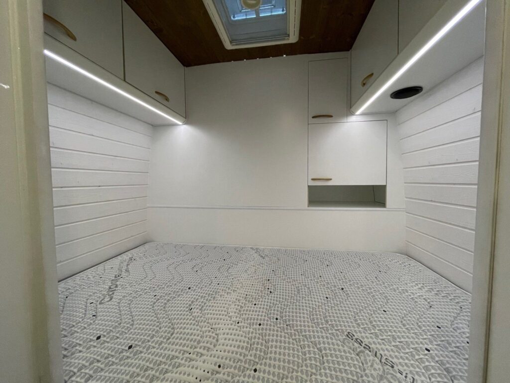 Interior de la Fiat Ducato L4H3 2+2 mostrando un área de dormitorio con una cama de matrimonio cubierta con un colchón de espuma con diseño geométrico, rodeado de paredes blancas y armarios superiores, con iluminación LED y una claraboya arriba.