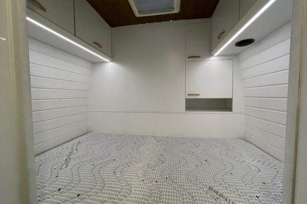 Interior de la Fiat Ducato L4H3 2+2 mostrando un área de dormitorio con una cama de matrimonio cubierta con un colchón de espuma con diseño geométrico, rodeado de paredes blancas y armarios superiores, con iluminación LED y una claraboya arriba.