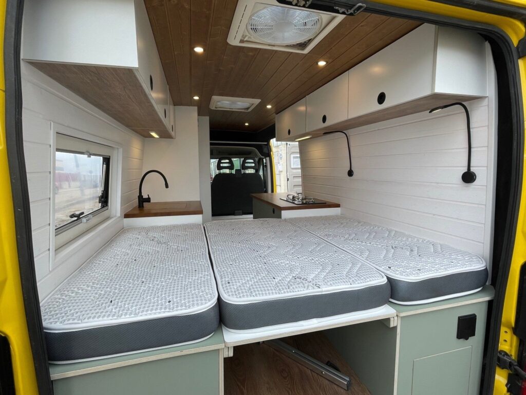Interior de la furgoneta camperizada Boxer L3H2 2+2 mostrando dos camas individuales con colchones de espuma con cubierta blanca, armarios superiores y una ventana lateral. El techo de madera y las paredes blancas crean un espacio acogedor y luminoso.