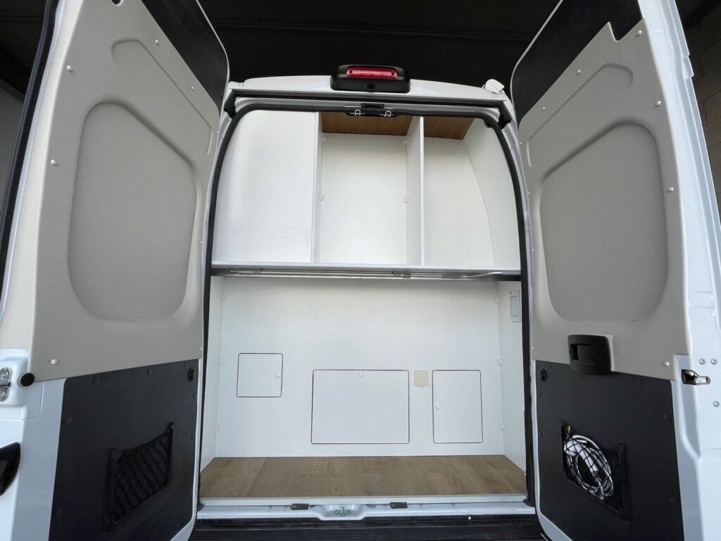 Vista del área de garaje en una Fiat Ducato L4H3 2+2, con las puertas traseras abiertas mostrando un espacio amplio y vacío listo para almacenamiento, con paredes y piso de color claro y una estructura de estante superior.