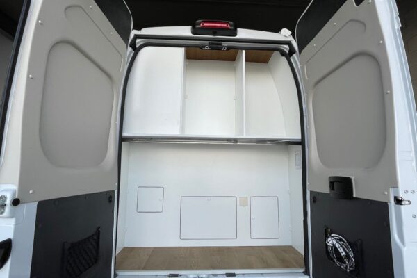 Vista del área de garaje en una Fiat Ducato L4H3 2+2, con las puertas traseras abiertas mostrando un espacio amplio y vacío listo para almacenamiento, con paredes y piso de color claro y una estructura de estante superior.