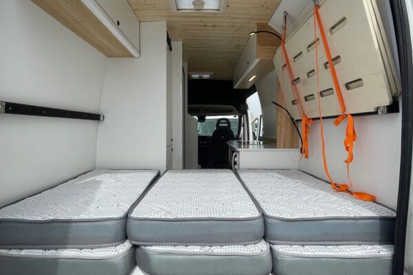 Detalle del espacio con los colchones sobre la cama principal, vista de atrás hacia delante de la Citroën Jumper L4H3 4+4, convertida por Baovan Camper, con acabado en blanco y techo de madera.