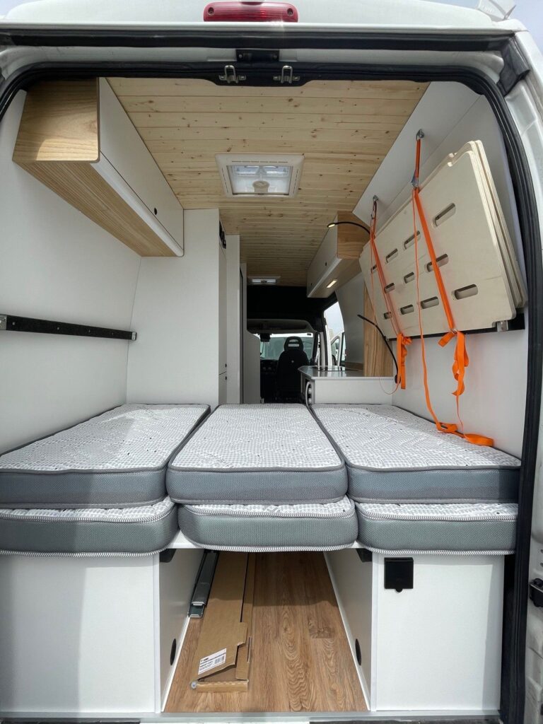 Detalle del espacio con los colchones sobre la cama principal, vista de atrás hacia delante de la Citroën Jumper L4H3 4+4, convertida por Baovan Camper, con acabado en blanco y techo de madera.