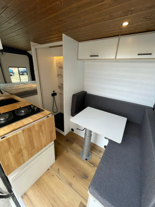 Área de descanso y comedor en la Camper L3H2 con asientos en 'L', mesa blanca ajustable y armarios superiores para almacenamiento eficiente.