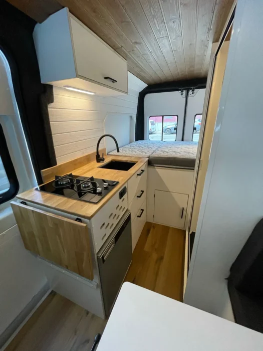 cocina funcional en la Camper L3H2 con estufa de gas, fregadero y encimeras de madera, diseñada para viajes cómodos.
