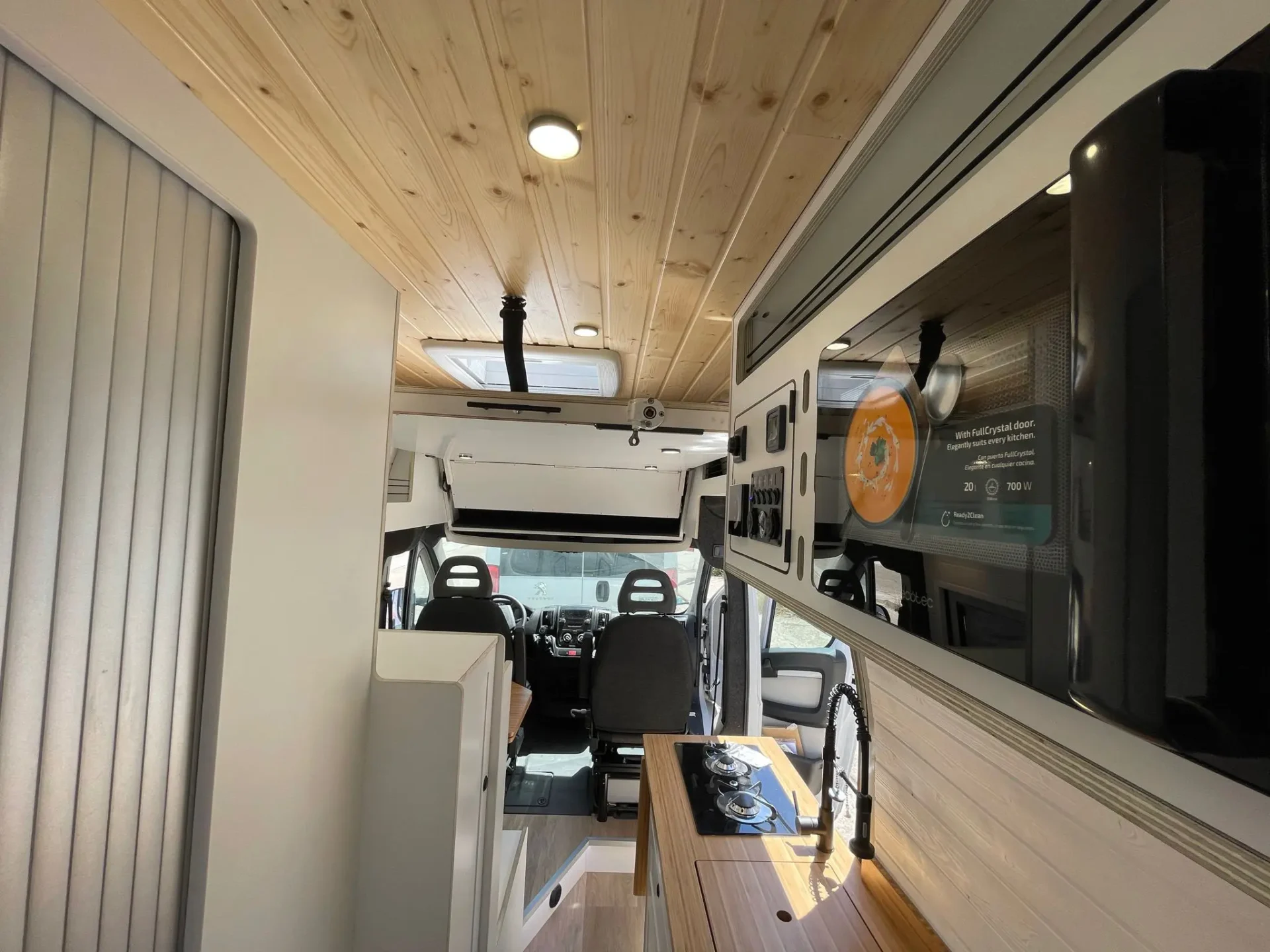 Vista de la zona de conducción desde el interior de la camperización de furgoneta Ducato L4H3 con cama abatible, destacando los acabados de madera en el techo y almacenamiento amplio.