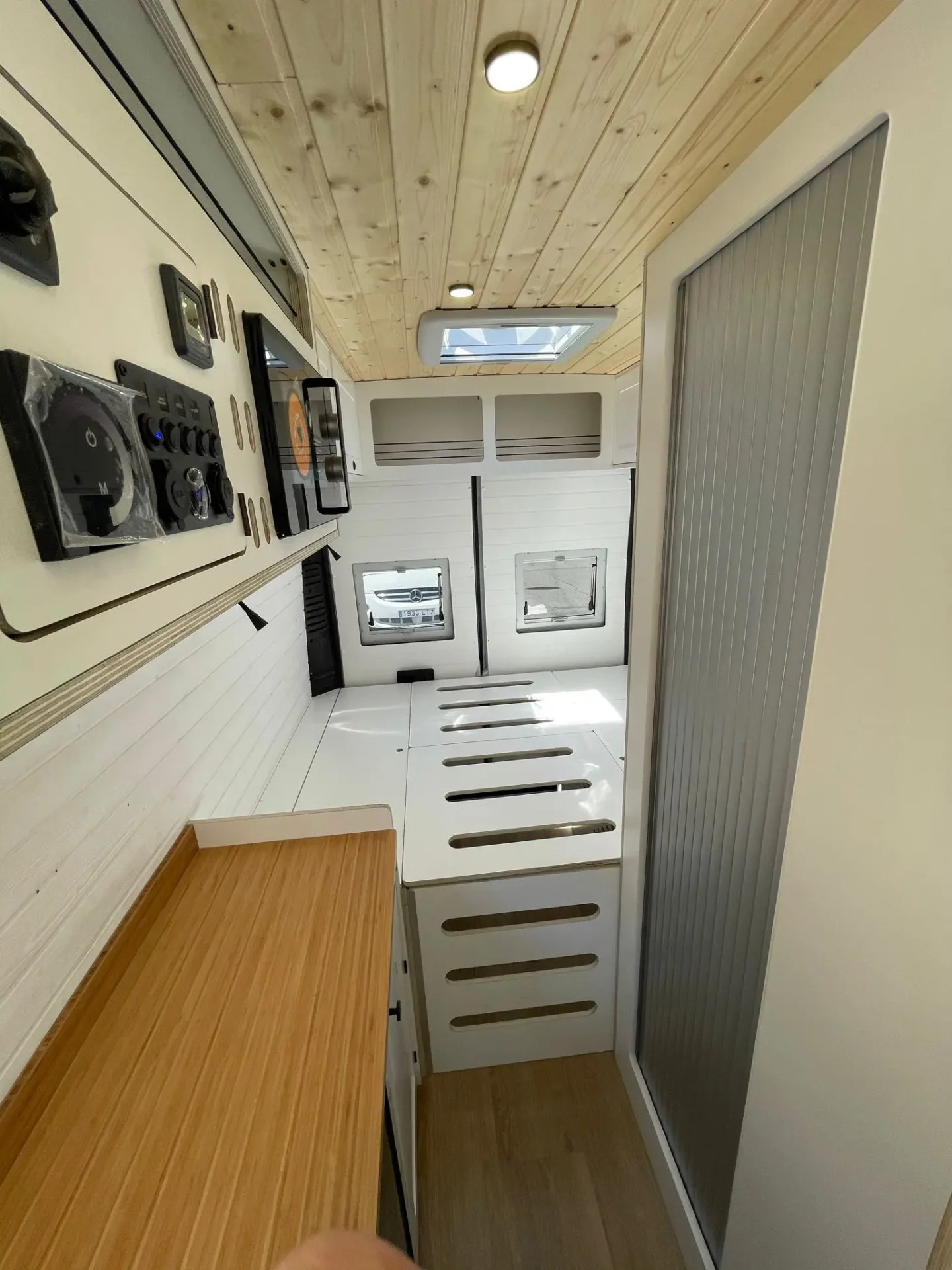 Interior moderno y minimalista de una furgoneta Ducato L4H3 camper con cama abatible sobre cabina, mostrando una cocina compacta con encimera de madera y gabinetes blancos.
