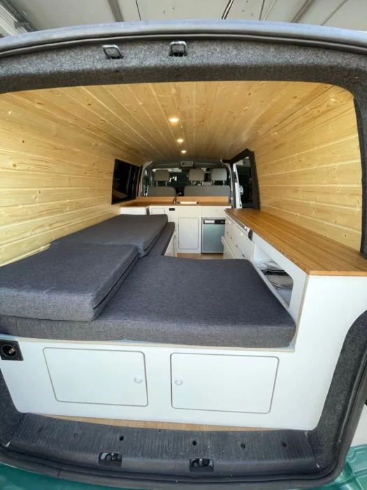 Vista desde la parte trasera de la VW T6 Camper hacia delante, mostrando la zona de cama confortable y el interior de madera con armarios blancos.