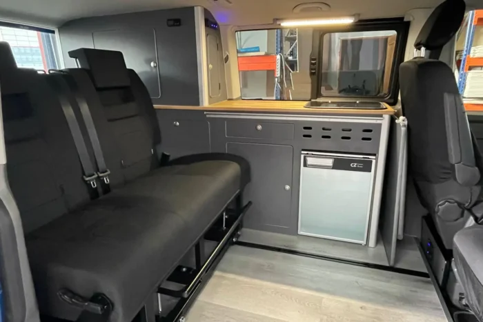 VW T6 camper con mueble lateral izquierdo y asiento convertible en cama corrediza, mostrando cocina integrada y almacenamiento