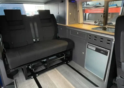 VW T6 Camper con detalles del espacio de descanso: sillas y mesa plegable plegada, nevera y espacio para cocinar.