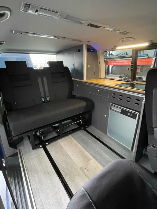 VW T6 Camper con detalles del espacio de descanso: sillas y mesa plegable plegada, nevera y espacio para cocinar.