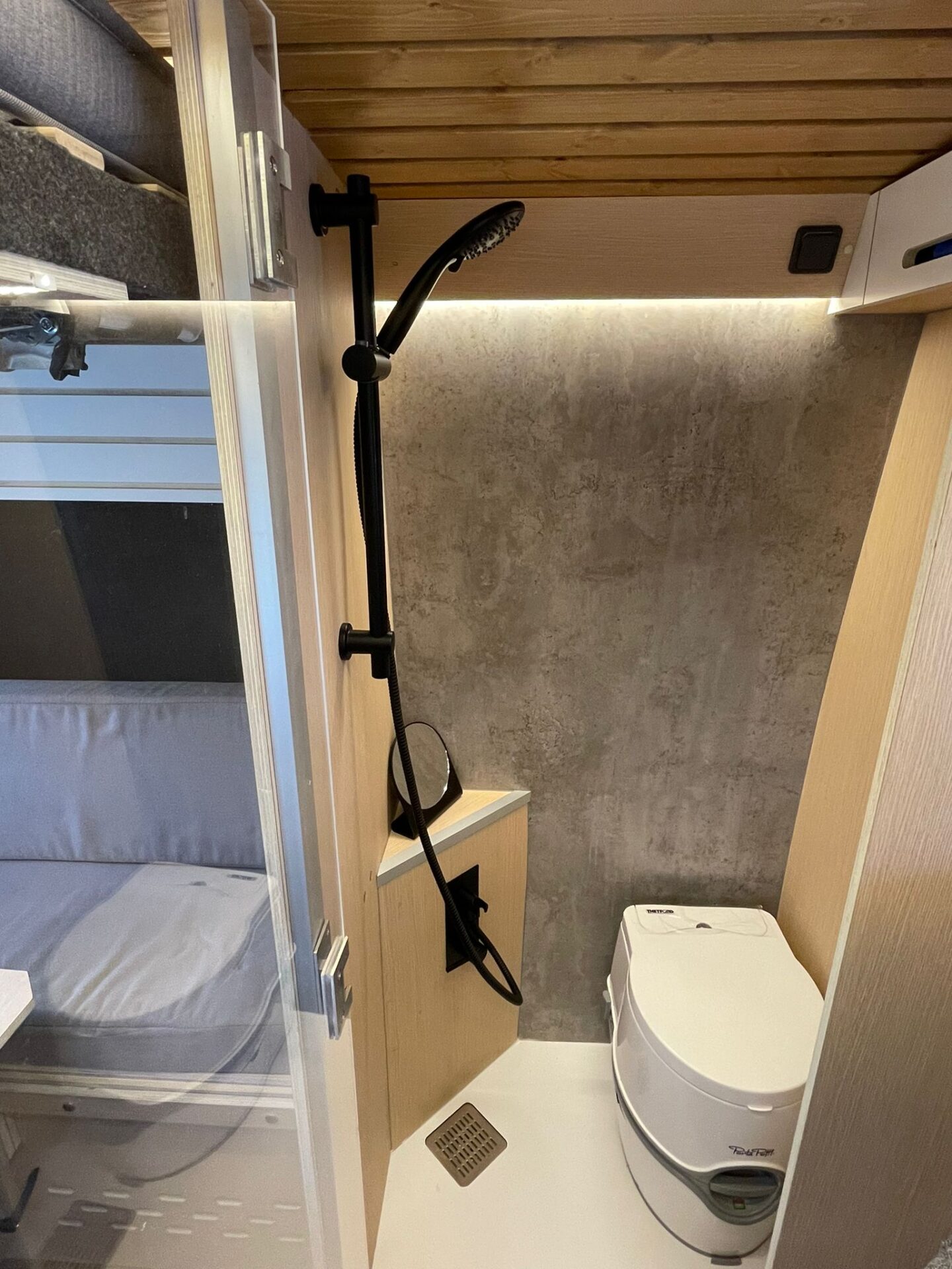 Interior del baño en camper Baovan con ducha negra ajustable y WC portátil Thetford, junto a la pared texturizada